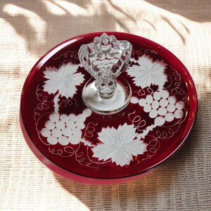 Può includere: Un vassoio da portata in vetro rosso con manico in vetro trasparente. Il vassoio presenta disegni di uva e foglie incisi in bianco. Il manico ha la forma di un grappolo d'uva. Il vassoio è rotondo e ha un bordo rialzato.