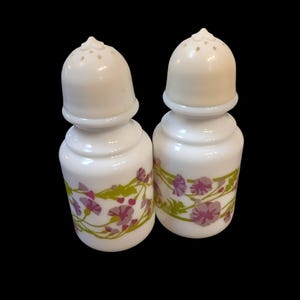 May include: A pair of white ceramic salt and pepper shakers with floral patterns. Each shaker has a rounded base and a cream-coloured, perforated lid. The floral design includes purple flowers and green leaves.