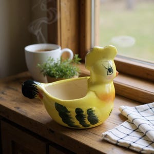 May include: A yellow ceramic chicken-shaped planter with black and green accents. The planter has a wide opening and a detailed face. A white mug with tea and a small plant are in the background. The planter is on a wooden surface.