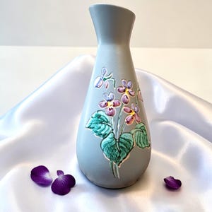 Puede incluir: Un jarrón de cerámica gris con un diseño floral de flores moradas y amarillas y hojas verdes. El jarrón tiene una forma curva y detalles en relieve. Pétalos de flores moradas dispersos cerca del jarrón.