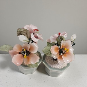 Op de afbeelding: Twee decoratieve bloemstukken met roze en perzikbloemen en groene bladeren. De bloemen staan in kleine, lichtblauwe zeshoekige potten gevuld met witte kiezelstenen. De bloemen hebben gele centra.