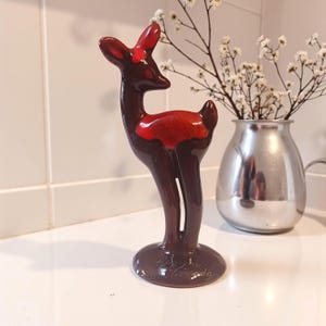 May include: A ceramic deer figurine in shades of brown and red. The deer has a slender body, a red bow, and a glossy finish. The figurine is standing on a round base. A silver vase with white flowers is in the background.