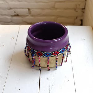 Könnte beinhalten: Ein Keramiktopf mit tiefviolettem Rand und beigem Körper. Der Topf ist mit einem farbenfrohen gewebten Band mit roten, blauen und weißen geometrischen Mustern verziert. Strähnen aus bunten Perlen hängen vom unteren Rand, was eine dekorative Note verleiht.