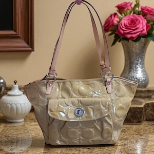 May include: A beige Coach tote bag with a glossy finish and a repeating logo pattern. The bag has light pink leather straps and a front pocket with a silver clasp. A vase of pink roses is in the background.