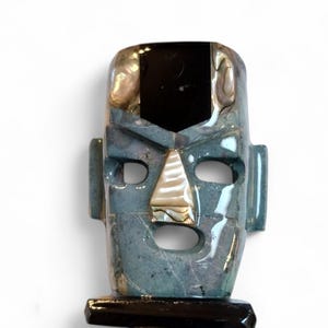 May include: A blue and black stone mask with a carved face and inlaid abalone shell details. The mask is on a black wooden base.