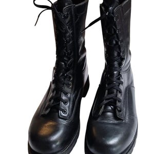 Puede incluir: Un par de botas de combate de cuero negro. Las botas están atadas en la parte delantera con cordones negros y tienen una punta redondeada. Las botas miden aproximadamente 25 cm de alto.