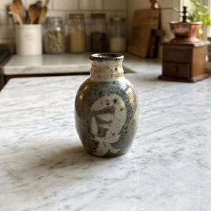 Peut inclure: Un petit vase en céramique fait à la main avec une finition tachetée et mouchetée dans des tons de marron, de gris et de crème. Le vase présente un motif abstrait unique sur sa surface. Le vase est exposé sur un comptoir en marbre.