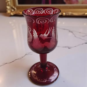 May include: A deep red glass with an etched design. The glass has a bird and foliage motif, with a swirl pattern around the rim and base. The stem is slender, and the base is round. A decorative glass.