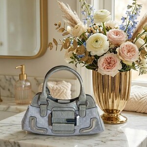 May include: A silver and grey handbag with a structured design rests on a marble surface. The bag has a top handle and a rectangular clasp. A gold vase filled with flowers is in the background.