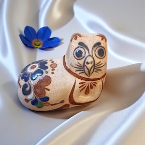 May include: A ceramic owl ornament with a beige body, decorated with brown and blue floral patterns. The owl features large eyes and a detailed beak. A blue flower with a yellow centre is placed nearby.