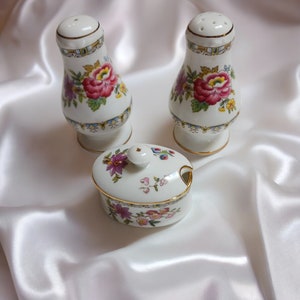 May include: A white porcelain salt and pepper shaker set and a small lidded dish. The set features a floral design with red, purple, and yellow flowers, accented with gold trim. The background is a white satin fabric.