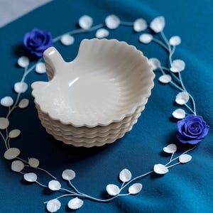 May include: A stack of cream-coloured, leaf-shaped ceramic dishes with a scalloped edge. The dishes are on a teal blue surface, decorated with a white floral garland and two blue rose accents. These dishes are suitable for serving appetizers or small portions.