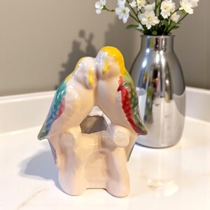 May include: A ceramic bird figurine with two parakeets kissing, perched on a light beige base. The birds have yellow heads, green and red wings, and a light beige body. A silver vase with white flowers is in the background.