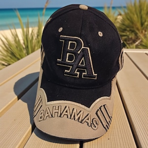 May include: A navy blue baseball cap with the letters "BA" embroidered in light tan. The word "BAHAMAS" is embroidered in light tan across the brim. The cap is sitting on a wooden surface with a beach and ocean in the background.