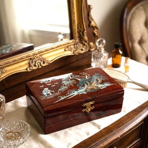 Può includere: Un portagioie rettangolare in legno scuro con un design intarsiato raffigurante un paesaggio con una figura, alberi e uccelli. La scatola ha una chiusura color oro ed è esposta su una superficie bianca.