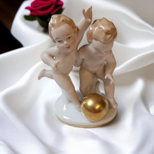 Könnte beinhalten: Porzellanfigur, die zwei cherubische Figuren zeigt, die mit einem goldenen Ball spielen. Die Figuren sind nackt mit detaillierten Gesichtszügen und stehen auf einem weißen Sockel mit goldenem Rand. Im Hintergrund ist eine Rose zu sehen.