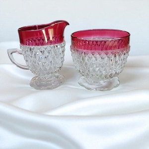 May include: A clear glass creamer and sugar bowl set with a ruby red rim. The creamer has a handle and spout. Both pieces have a diamond pattern and sit on a clear, scalloped base. The set is displayed on a white, silky surface.