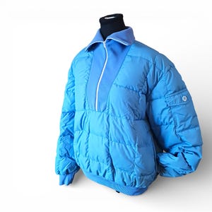 Puede incluir: Chaqueta acolchada azul claro con cierre de media cremallera y cuello azul claro en contraste. La chaqueta tiene un pequeño bolsillo en la manga y se exhibe sobre una cabeza de maniquí negra. La chaqueta es de estilo abullonado.
