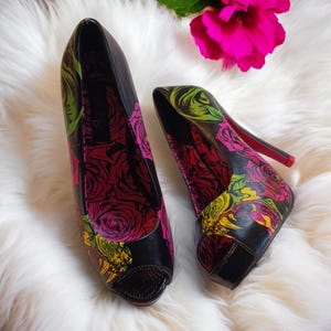 May include: Pair of black peep-toe high heels with a vibrant floral pattern in pink, yellow, and green. The heels have a red sole and are set against a white, fluffy background.