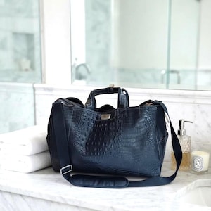 May include: A dark navy blue duffel bag with a textured crocodile skin pattern. The bag features two top handles, a long adjustable shoulder strap with a padded section, and a small rectangular silver-toned metal plate on the front centre.