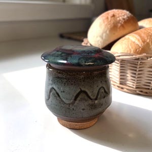 Puede incluir: Una mantequera de cerámica con tapa. El plato es gris moteado con un diseño ondulado, y la tapa es de color verde oscuro y rojo. El plato se asienta sobre una base de corcho. Una cesta de panecillos está en el fondo.