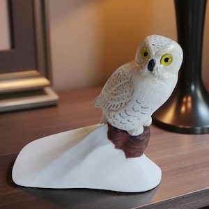 Puede incluir: Figura de búho de cerámica blanca con ojos amarillos y pico negro, posada sobre una base marrón y blanca. El búho tiene plumas texturizadas. Es un objeto decorativo.