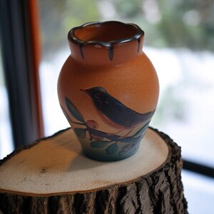 Peut inclure: Petit vase en céramique orange avec un oiseau peint sur le devant. Le vase a un bord festonné et est posé sur une tranche de bois. L'oiseau est noir avec une poitrine rouge, perché sur une branche avec des feuilles et des baies.
