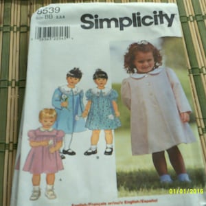 May include: Simplicity sewing pattern number 8539 for girls' dresses. The pattern includes three different dress styles: a blue dress with a white collar, a blue and white polka dot dress, and a pink dress with a white collar. The pattern also includes a white dress with a white collar and long sleeves.