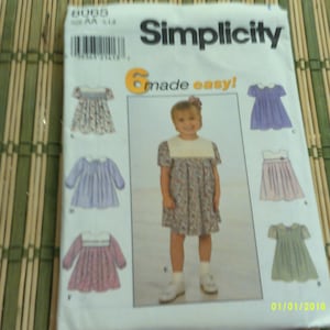 May include: Simplicity sewing pattern for girls' dresses. The pattern features multiple dress designs, including options with long and short sleeves, and various collar styles. The package includes a photo of a young girl wearing one of the dresses.