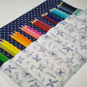 May include: A fabric pencil roll with a navy blue polka dot exterior and a white interior patterned with blue crosses and inspirational words. The roll contains a variety of colored pencils, arranged by color.
