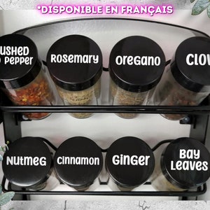 Peut inclure: Un porte-épices en métal noir avec huit bocaux en verre contenant diverses épices. Les bocaux ont des couvercles noirs avec des étiquettes blanches qui indiquent "poivre rouge moulu", "romarin", "origan", "clou de girofle", "muscade", "cannelle", "gingembre" et "feuilles de laurier".