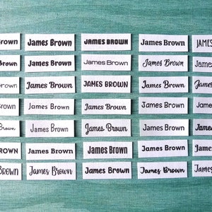 Op de afbeelding: Vierentwintig witte rechthoekige labels met de tekst "James Brown" in zwart gedrukt. De labels zijn gerangschikt in zes rijen van vier labels.
