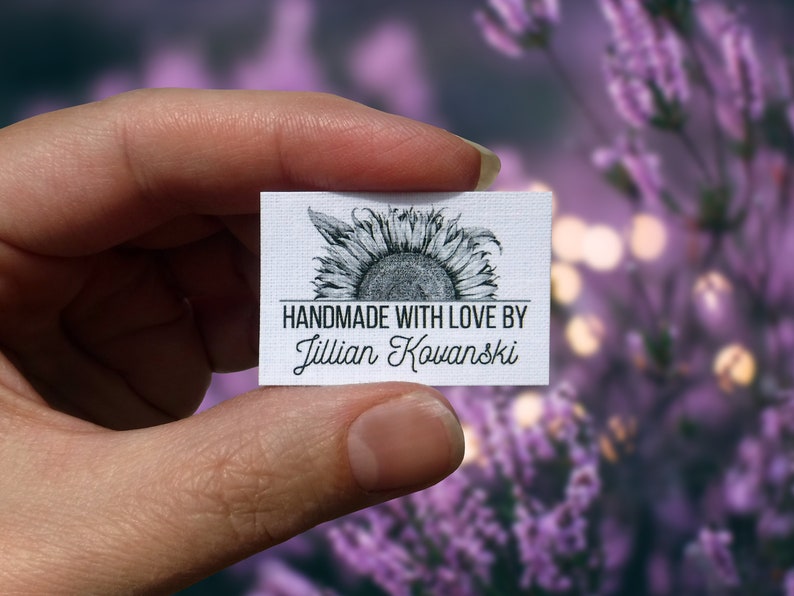 Pode incluir: Um r&oacute;tulo de tecido branco com um gr&aacute;fico de girassol preto e branco. O texto "HANDMADE WITH LOVE BY Jillian Kowanski" &eacute; impresso abaixo do girassol.