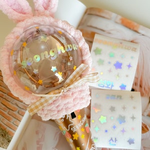 May include: A pink fluffy bunny ear headband with a clear plastic sphere decorated with iridescent stars and the word "Seonghwa" in gold lettering. The sphere is attached to a gold-colored light stick with a checkered pattern and the word "Seonghwa" repeated on the stick. The light stick is decorated with iridescent stars and strawberries.