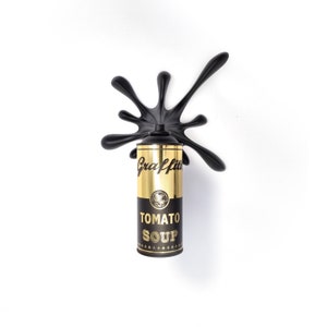 May include: A gold can of tomato soup with the word "Graffiti" written in black cursive on the front. The can is covered in a black paint splatter.