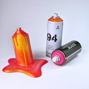 Puede incluir: Composición artística con tres botes de pintura en aerosol. Un bote naranja y amarillo, con pintura goteando. Un bote blanco con "94" y un bote negro con pintura rosa también están presentes.