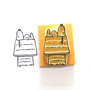 May include: A Snoopy-themed rubber stamp featuring Snoopy lounging on top of his doghouse. The stamp is mounted on a rectangular wooden block. A black and white line drawing of the same image is next to the stamp.