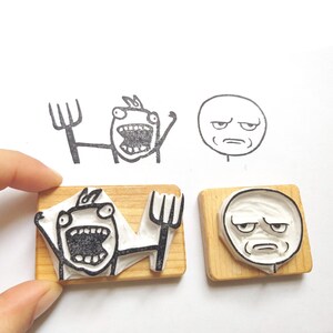 May include: Two wooden blocks with rubber stamps featuring different internet meme faces. One stamp shows a person with a surprised expression and the other shows a person with a confused expression.