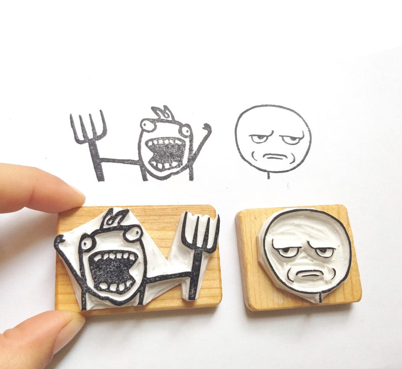 May include: Two wooden blocks with rubber stamps featuring different internet meme faces. One stamp shows a person with a surprised expression and the other shows a person with a confused expression.