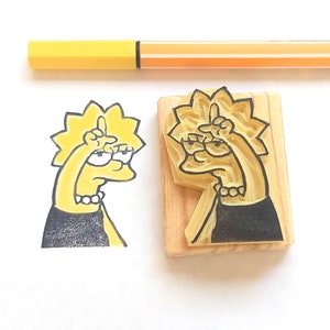 Puede incluir: Un sello de goma amarillo y negro de un personaje de dibujos animados con la mano levantada, junto a su imagen estampada. El sello está montado sobre un bloque de madera. Un bolígrafo amarillo y naranja está en el fondo. La imagen estampada es un personaje de dibujos animados.
