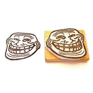 May include: A wooden rubber stamp with a black and white image of the troll face meme. The troll face is smiling with a mischievous grin and has one eyebrow raised.