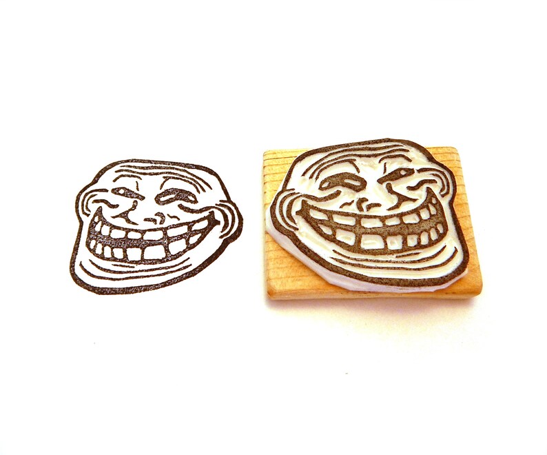 May include: A wooden rubber stamp with a black and white image of the troll face meme. The troll face is smiling with a mischievous grin and has one eyebrow raised.