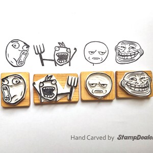 May include: Six wooden stamps featuring various internet meme faces, including Trollface,  Forever Alone, and  'You're doing it wrong'. The stamps are hand-carved and have a light brown color. The text 'Hand Carved by StampDealer' is printed below the stamps.