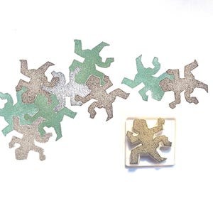 Puede incluir: Una colección de figuras humanas abstractas y brillantes en plata, verde y bronceado. También hay un sello cuadrado transparente con una figura a juego. Las figuras están dispersas sobre un fondo blanco.