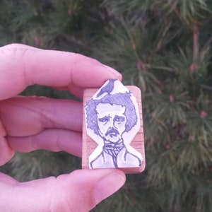 May include: A wooden block stamp with a black and white illustration of Edgar Allan Poe. The illustration shows Poe with a dark beard and mustache, wearing a dark shirt and a white collar.