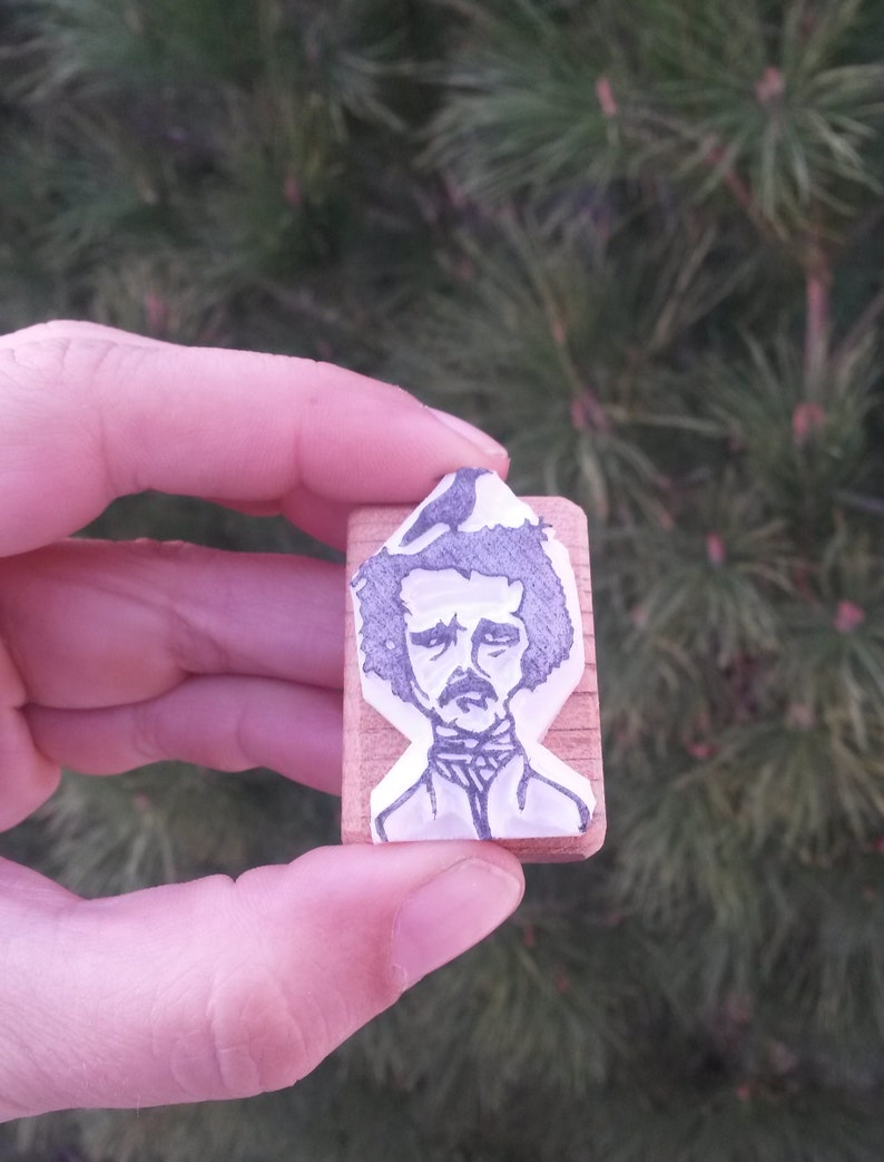 May include: A wooden block stamp with a black and white illustration of Edgar Allan Poe. The illustration shows Poe with a dark beard and mustache, wearing a dark shirt and a white collar.