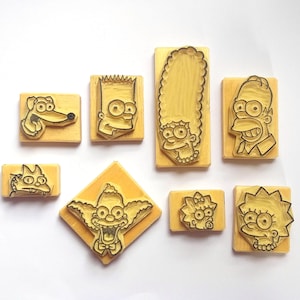 May include: A collection of eight yellow rubber stamps featuring cartoon character designs. The stamps include images of a dog, Bart Simpson, Marge Simpson, Homer Simpson, Krusty the Clown, and Lisa Simpson. The stamps are mounted on wooden blocks.