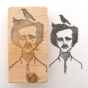 May include: A wooden stamp with a black and white illustration of Edgar Allan Poe with a bird perched on his head. The stamp is ready to be used to create a unique design.