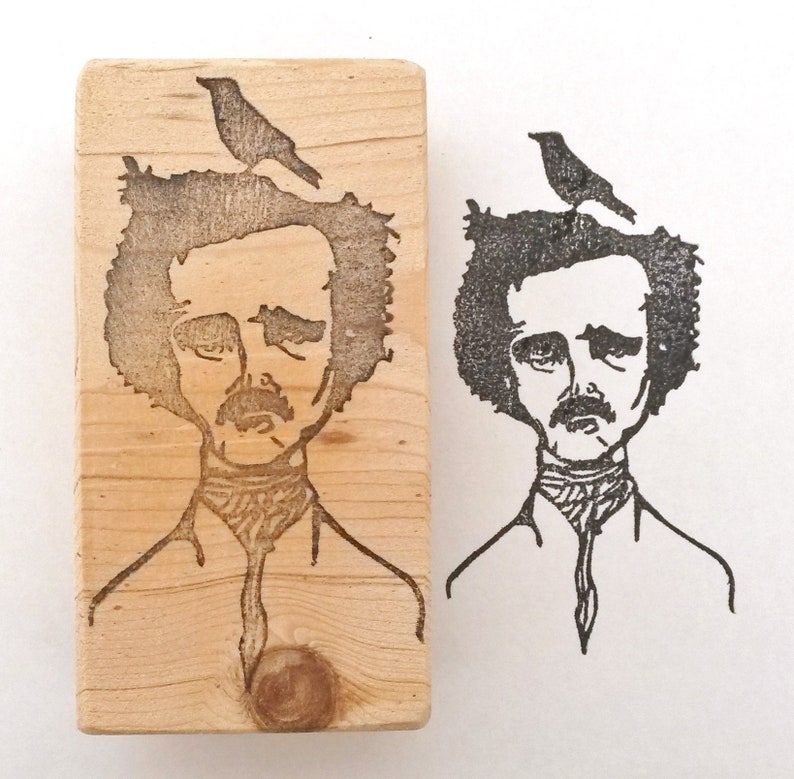 May include: A wooden stamp with a black and white illustration of Edgar Allan Poe with a bird perched on his head. The stamp is ready to be used to create a unique design.