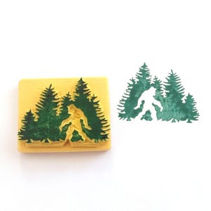 Puede incluir: Un sello de goma de madera con una silueta verde de Bigfoot caminando por un bosque de pinos.